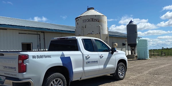 A white truck with the New Standard logo is parked outside a warehouse or farm. The scene features a clear, beautiful sky, with some grass in the middle of the photo. A few tank-like containers are visible near the truck, positioned in front of the warehouse.