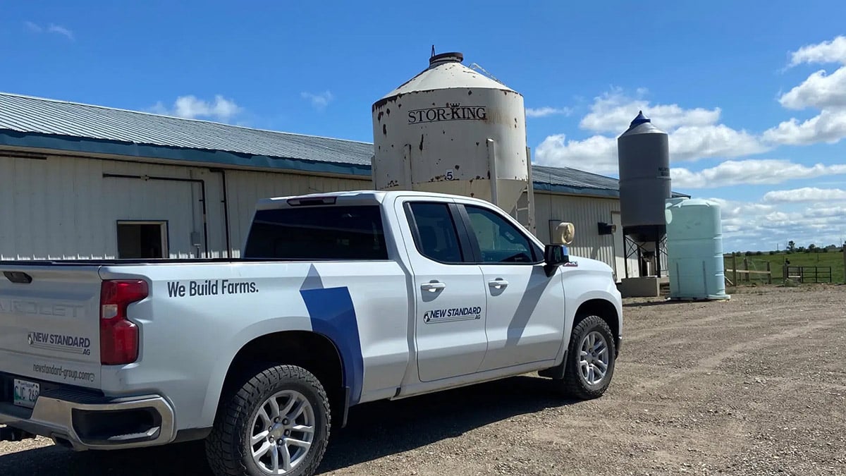 Pickup truck with 'We Build Farms' and 'New Standard AG' branding parked beside farm silos and a barn under a clear blue sky